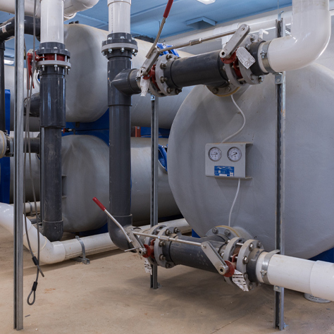 Pictured is a series of stacked and single fiberglass commercial pool filters in a pool filter room. Also shown are the face piping, plumbing and valves that are connected.