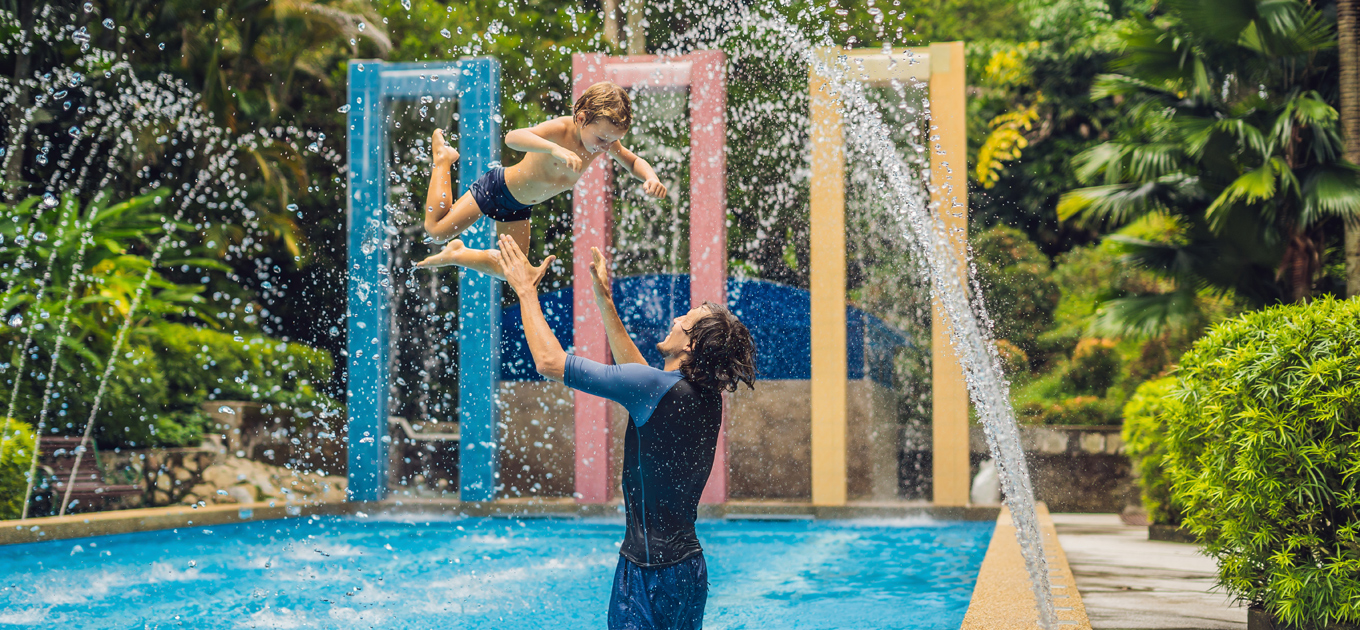 Images shows a dad tossing his son up in the air and catching him in a waterpark pool with water fountains spraying water while they play in the pool.