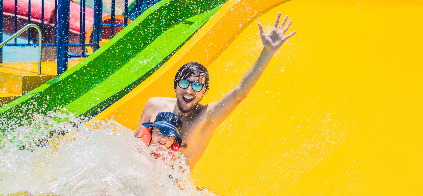 Image shows a man with sunglasses and a beard waving as he goes down a yellow waterslide at a water park with a small child with visor in his lap.