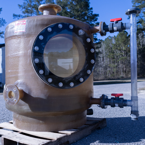 This is an image of a Mer-Made Filter fiberglass commercial pool surge tank on a wooden pallet. The tank is tan-colored fiberglass with an outlet on the lower left side and a large manway with clear cover. The right side has two outlets that connect to a clear tube with shut off valves at each end.